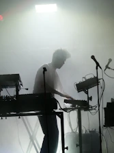 A silhouette of a person engaged in a live electronic music performance, surrounded by various sound equipment such as keyboards, synthesizers, and microphones. The stage is filled with fog, creating a moody and atmospheric ambiance. A strong light from above adds to the dramatic effect.
