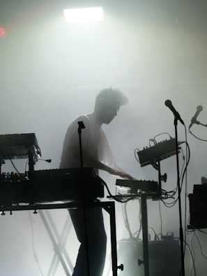 A silhouette of a person engaged in a live electronic music performance, surrounded by various sound equipment such as keyboards, synthesizers, and microphones. The stage is filled with fog, creating a moody and atmospheric ambiance. A strong light from above adds to the dramatic effect.