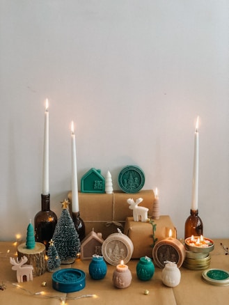 A collection of Christmas-themed decorative items are arranged on a table. Several tall, white candles are burning in brown glass bottles. There are small, artificial Christmas trees, a miniature white ceramic moose, and various ornament-like candles in teal, white, and cream shades. The backdrop features wrapped gifts tied with string, and there are wooden tree slices used as a base for other decorations. Small fairy lights add a warm glow to the scene.