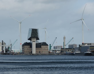 A waterfront area featuring a modern building with a unique glass structure on top of an older architectural base. Several large wind turbines are visible in the background, along with cranes and industrial structures.
