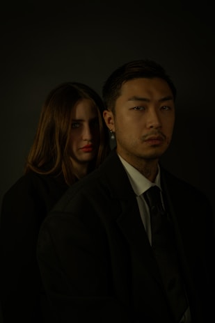 A man and a woman dressed in dark, formal attire. The ambient lighting is dim, creating a mysterious and moody atmosphere. The man is in the foreground with a serious expression, while the woman stands slightly behind him, gazing forward.