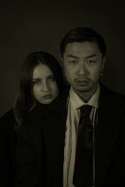 Two individuals, a woman and a man, are wearing formal attire, with the man standing slightly in front. They both have serious expressions on their faces. The lighting is dim, creating a dramatic and somber atmosphere.