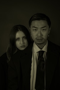Two individuals, a woman and a man, are wearing formal attire, with the man standing slightly in front. They both have serious expressions on their faces. The lighting is dim, creating a dramatic and somber atmosphere.