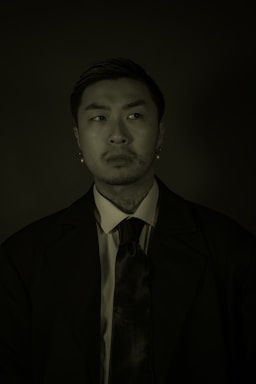 A man dressed in formal attire, including a suit and tie, is captured against a dark background. The lighting contrasts sharply, casting shadows over his face, suggesting a serious or contemplative expression.