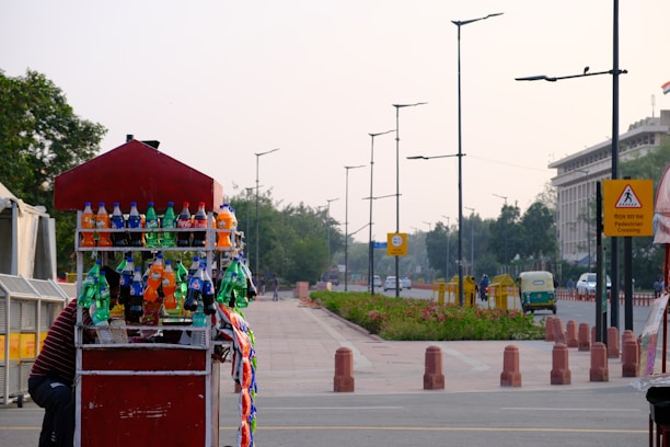 A roadside stall selling various bottled soft drinks is displayed prominently in the foreground, with colorful bottles like Sprite, Fanta, and Coca-Cola arranged on shelves. A pedestrian crossing sign is visible on the side of the road which is lined with trees and bushes. Vehicles including a green auto-rickshaw are seen moving on the road further ahead, with lampposts lining the pavement.