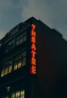 A theater building with large windows and a vintage architectural style. The neon sign spelling 'THEATRE' glows in bright red, creating a stark contrast against the dark facade. The upper part of the building displays the year '1912'. The sky appears overcast, adding a moody atmosphere.
