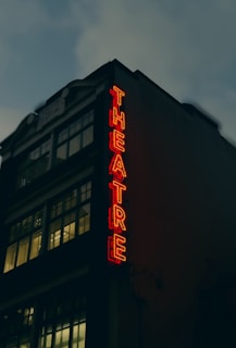 A theater building with large windows and a vintage architectural style. The neon sign spelling 'THEATRE' glows in bright red, creating a stark contrast against the dark facade. The upper part of the building displays the year '1912'. The sky appears overcast, adding a moody atmosphere.