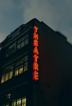 A theater building with large windows and a vintage architectural style. The neon sign spelling 'THEATRE' glows in bright red, creating a stark contrast against the dark facade. The upper part of the building displays the year '1912'. The sky appears overcast, adding a moody atmosphere.