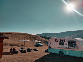 A coffee trailer is set up on an open field under a clear blue sky. There are two white umbrellas with outdoor seating, and several people are seated nearby on the ground. A car is parked in the background with rolling hills surrounding the area. The sun shines brightly, casting shadows across the scene.