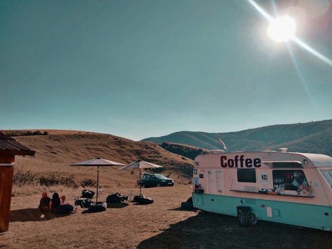 A coffee trailer is set up on an open field under a clear blue sky. There are two white umbrellas with outdoor seating, and several people are seated nearby on the ground. A car is parked in the background with rolling hills surrounding the area. The sun shines brightly, casting shadows across the scene.