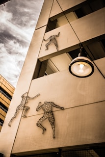 Metal sculptures of human figures are mounted on the exterior of a building, appearing to be climbing up the structure. The building has a modern architectural style with visible beams and a nearby streetlamp. The sky is partly cloudy, adding a dynamic background to the scene.