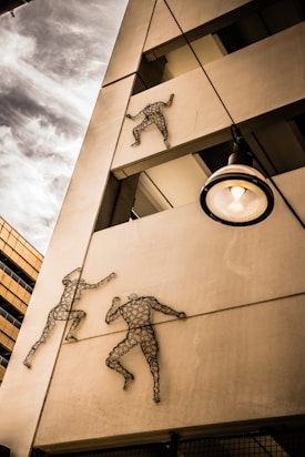Metal sculptures of human figures are mounted on the exterior of a building, appearing to be climbing up the structure. The building has a modern architectural style with visible beams and a nearby streetlamp. The sky is partly cloudy, adding a dynamic background to the scene.
