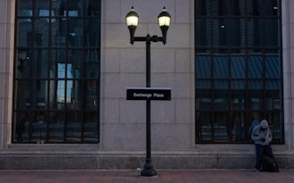 A building facade with large grid-patterned windows and a signpost indicating 'Exchange Place'. A person in a hoodie stands to the right, leaning against the wall, looking at their phone under dim lighting provided by two street lamps.