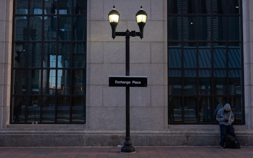 A building facade with large grid-patterned windows and a signpost indicating 'Exchange Place'. A person in a hoodie stands to the right, leaning against the wall, looking at their phone under dim lighting provided by two street lamps.