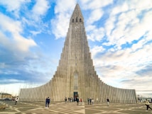 A towering concrete church with a striking, modern design featuring a tall, central spire. It stands against a partly cloudy blue sky, surrounded by a few people walking and taking photos in the plaza in front of it.