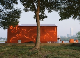A vibrant red structure is adorned with fluid, organic reflections that appear like fiery accents. In the foreground, a tree with lush green leaves casts a shadow over the scene. Several transparent bottles and orange crates are arranged near the building. The background features a clear sky and distant cityscape, adding a serene ambiance.