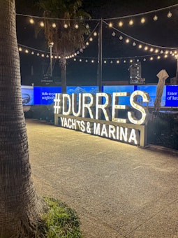 A nighttime scene featuring a brightly lit sign that reads '#DURRES YACHTS & MARINA.' The atmosphere is festive with strings of lights hanging above, casting a warm glow over the area. Palm trees and a paved pathway are visible in the foreground. The background includes illuminated advertising screens and structures.