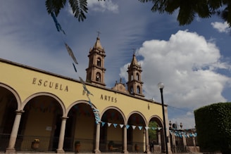 An architectural structure with arched columns and a facade that reads 'Escuela de Artes'. Two bell towers rise into a partly cloudy sky. Blue and green decorative flags hang across the foreground, and the area is surrounded by greenery.