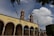 An architectural structure with arched columns and a facade that reads 'Escuela de Artes'. Two bell towers rise into a partly cloudy sky. Blue and green decorative flags hang across the foreground, and the area is surrounded by greenery.