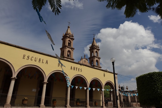 An architectural structure with arched columns and a facade that reads 'Escuela de Artes'. Two bell towers rise into a partly cloudy sky. Blue and green decorative flags hang across the foreground, and the area is surrounded by greenery.