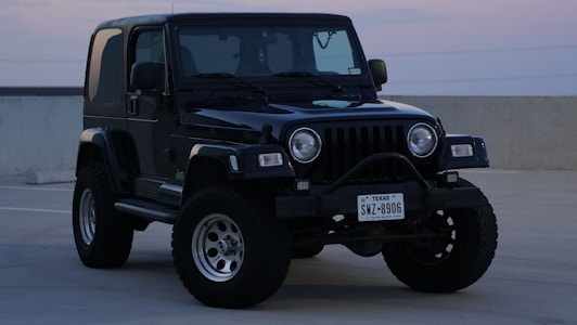 A black off-road vehicle, likely a Jeep, is parked on a concrete surface in an exterior setting. It features large tires, a rugged front bumper, and a visible Texas license plate. The scene appears to be captured during dusk or dawn, with soft lighting.