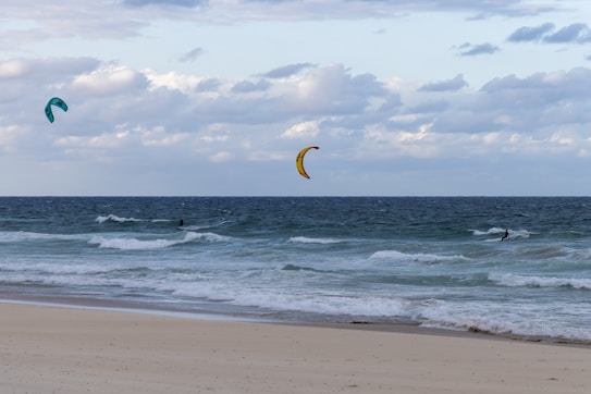 Kitesurfers ride the waves near the shore, with colorful kites in the sky above a blue ocean. The beach is sandy and appears calm, while the sky is cloudy, adding a moody atmosphere.