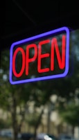 A neon sign with the word 'OPEN' in bright red letters, surrounded by a glowing blue border, is visible. The sign appears to be in a window, with blurred trees and buildings in the background, indicating an outdoor setting.