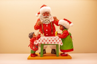 Santa Claus and two children are gathered around a table with a festive red and white polka dot tablecloth. Santa is wearing his traditional red suit and hat, and is holding a white mug. The children, dressed in festive red and green clothing, appear to be engaged in a holiday activity with Santa, one holding a Christmas decoration.