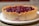 A round dessert featuring a crumbly, golden-brown crust filled with a rich, glossy cherry filling sits on a white plate. The crust is thick and has a slightly textured appearance, while the filling is vibrant and deeply colored, suggesting a freshly baked treat.