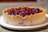 A round dessert featuring a crumbly, golden-brown crust filled with a rich, glossy cherry filling sits on a white plate. The crust is thick and has a slightly textured appearance, while the filling is vibrant and deeply colored, suggesting a freshly baked treat.