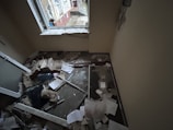 A small room with significant damage, including debris and papers scattered on the floor. The window frame appears broken, and insulation materials are exposed. There is a mix of building materials, like broken glass and wood, along with cardboard boxes.