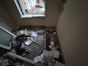 A small room with significant damage, including debris and papers scattered on the floor. The window frame appears broken, and insulation materials are exposed. There is a mix of building materials, like broken glass and wood, along with cardboard boxes.