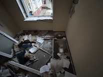 A small room with significant damage, including debris and papers scattered on the floor. The window frame appears broken, and insulation materials are exposed. There is a mix of building materials, like broken glass and wood, along with cardboard boxes.