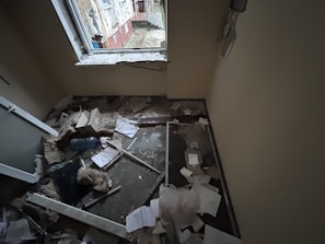 A small room with significant damage, including debris and papers scattered on the floor. The window frame appears broken, and insulation materials are exposed. There is a mix of building materials, like broken glass and wood, along with cardboard boxes.