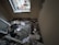 A small room with significant damage, including debris and papers scattered on the floor. The window frame appears broken, and insulation materials are exposed. There is a mix of building materials, like broken glass and wood, along with cardboard boxes.