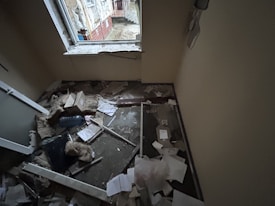 A small room with significant damage, including debris and papers scattered on the floor. The window frame appears broken, and insulation materials are exposed. There is a mix of building materials, like broken glass and wood, along with cardboard boxes.