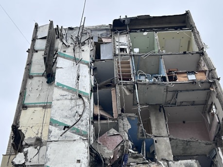 A heavily damaged building with exposed interior rooms and walls. The structure is partially collapsed, with debris hanging precariously. The floors and ceilings are barely intact, revealing furniture and objects inside. Electrical wires and pipes are visible among the debris.