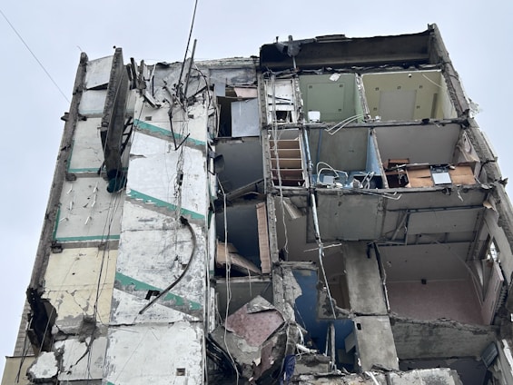A heavily damaged building with exposed interior rooms and walls. The structure is partially collapsed, with debris hanging precariously. The floors and ceilings are barely intact, revealing furniture and objects inside. Electrical wires and pipes are visible among the debris.