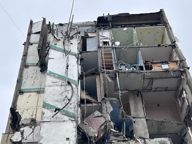 A heavily damaged building with exposed interior rooms and walls. The structure is partially collapsed, with debris hanging precariously. The floors and ceilings are barely intact, revealing furniture and objects inside. Electrical wires and pipes are visible among the debris.
