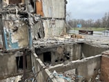 A heavily damaged building with significant structural destruction, showing exposed interiors and debris scattered throughout. The walls are partially collapsed, revealing electrical wires, pipes, and indistinguishable remains. The scene is outdoors, with a sparse, overcast sky and a deserted roadway visible in the background.