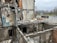 A heavily damaged building with significant structural destruction, showing exposed interiors and debris scattered throughout. The walls are partially collapsed, revealing electrical wires, pipes, and indistinguishable remains. The scene is outdoors, with a sparse, overcast sky and a deserted roadway visible in the background.