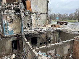 A heavily damaged building with significant structural destruction, showing exposed interiors and debris scattered throughout. The walls are partially collapsed, revealing electrical wires, pipes, and indistinguishable remains. The scene is outdoors, with a sparse, overcast sky and a deserted roadway visible in the background.
