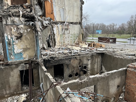 A heavily damaged building with significant structural destruction, showing exposed interiors and debris scattered throughout. The walls are partially collapsed, revealing electrical wires, pipes, and indistinguishable remains. The scene is outdoors, with a sparse, overcast sky and a deserted roadway visible in the background.