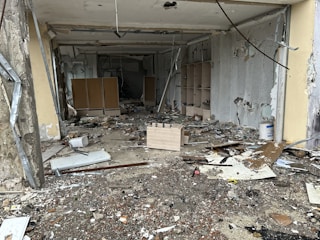 A room in disrepair with debris and broken items scattered across the floor. The walls are partially torn and damaged, with exposed cement and peeling paint. Wooden shelves and damaged furniture are visible towards the back.