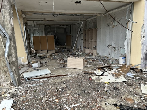 A room in disrepair with debris and broken items scattered across the floor. The walls are partially torn and damaged, with exposed cement and peeling paint. Wooden shelves and damaged furniture are visible towards the back.