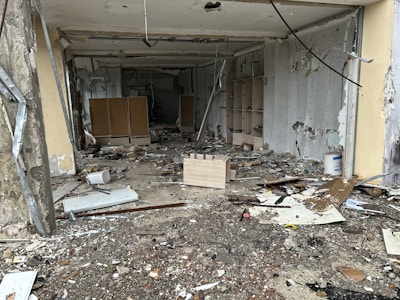 A room in disrepair with debris and broken items scattered across the floor. The walls are partially torn and damaged, with exposed cement and peeling paint. Wooden shelves and damaged furniture are visible towards the back.