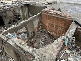 A partially demolished building showing exposed brick and concrete walls. Debris, including metal rods and wires, is scattered around the area. Holes can be seen in the walls, and some structures are broken down. There is a contrast between the reddish and grayish tones of the bricks and concrete.