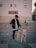 A person standing outdoors holding the leash of a white and tan dog. The setting appears to be an urban environment with paved ground and a building in the background. The person is wearing sunglasses and a black jacket with red accents.