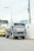A large gray truck with a tanker attached is parked on a road next to a wall with safety signage. A yellow truck is visible behind it. Overhead, multiple power lines are strung between utility poles.