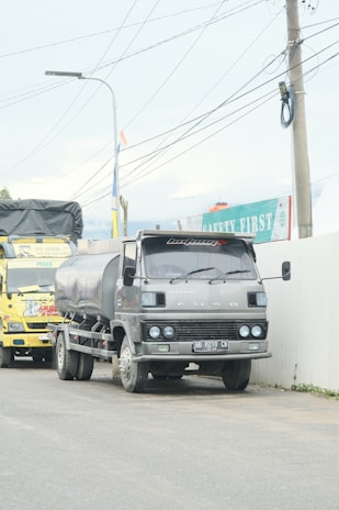 A large gray truck with a tanker attached is parked on a road next to a wall with safety signage. A yellow truck is visible behind it. Overhead, multiple power lines are strung between utility poles.